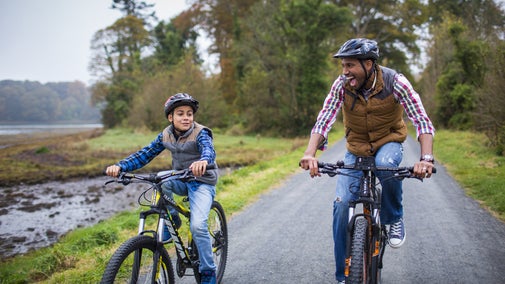 A man and his son cycle along the trail at Castle Ward, next to the shores of Strangford Lough. They're looking at each other, laughing and having fun as they cycle along.